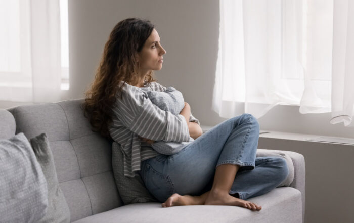 Woman sitting on a couch, looking pensively out a large window. She rests her chin on her hand, with trees visible outside.