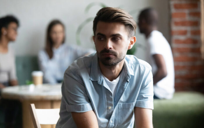 Man in denim shirt looking away, seated in a cafe. Two pairs of people converse in the blurred background.