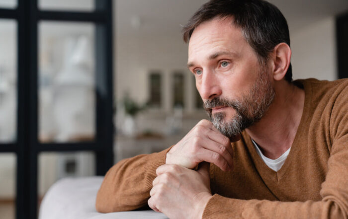 A man with a brown sweater looks pensively into the distance, resting his chin on his hand, in a softly lit interior space.