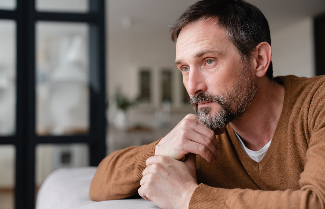 shutterstock_1998724388 A man with a brown sweater looks pensively into the distance, resting his chin on his hand, in a softly lit interior space.