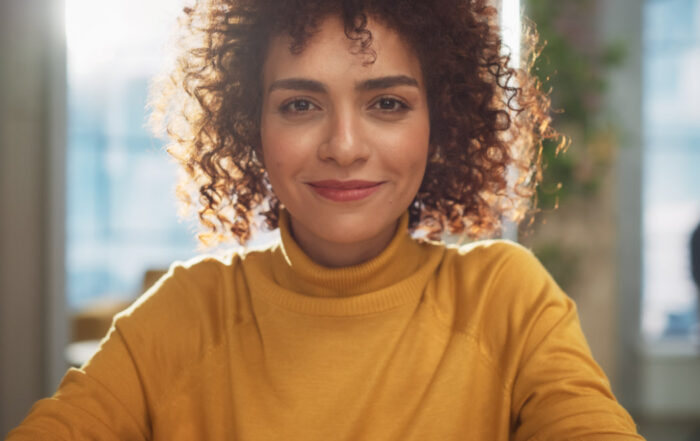 A person with curly hair and a yellow turtleneck sweater smiles while sitting indoors with sunlight in the background.