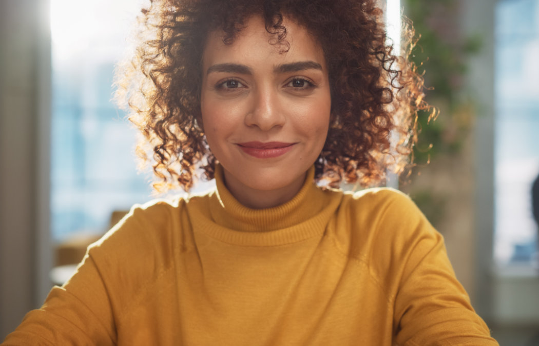 shutterstock_2231716615 A person with curly hair and a yellow turtleneck sweater smiles while sitting indoors with sunlight in the background.