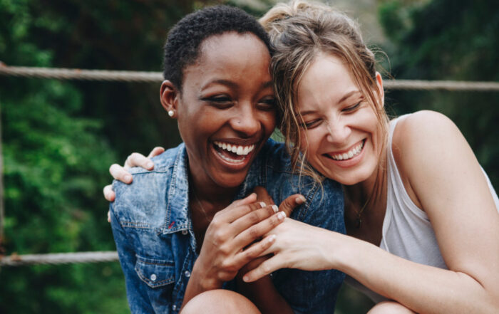 Two women smiling and laughing together outdoors, sitting closely with arms around each other. Green foliage is visible in the background.