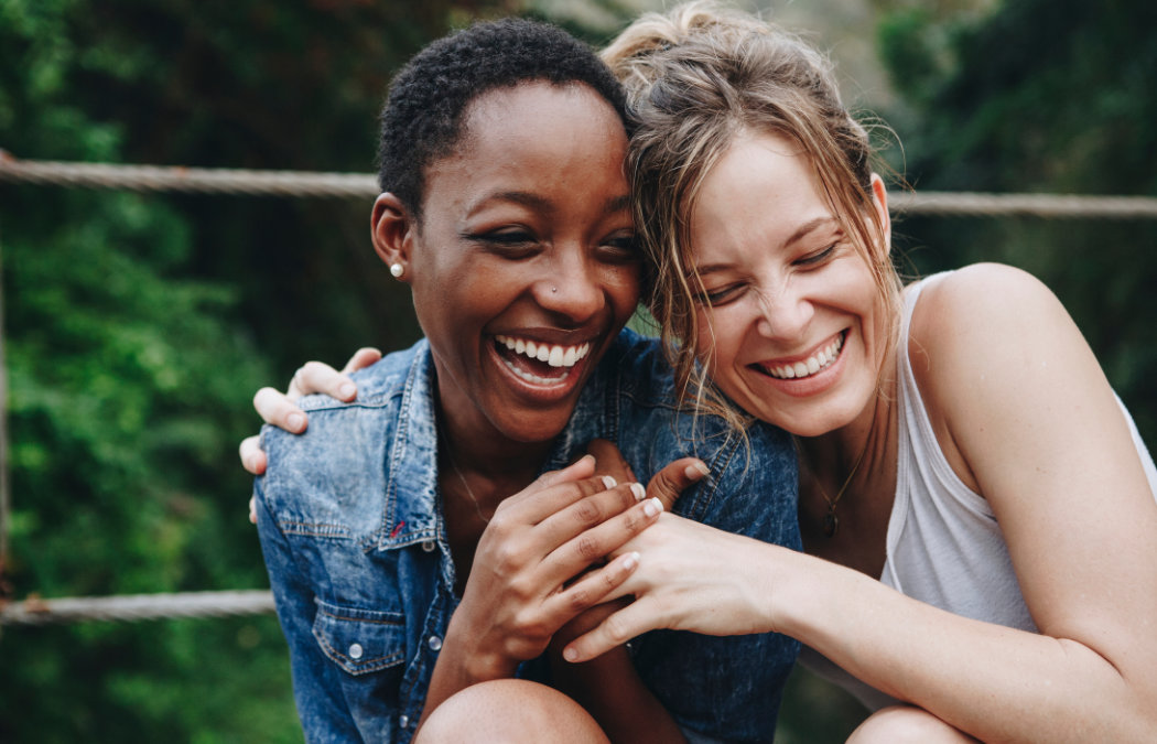 shutterstock_1038614926 Two women smiling and laughing together outdoors, sitting closely with arms around each other. Green foliage is visible in the background.