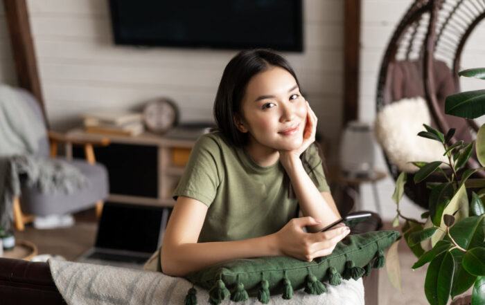 A young woman in a green t-shirt sits indoors, leaning on a cushion, holding a smartphone, and looking at the camera. A laptop and home decor are visible in the background.