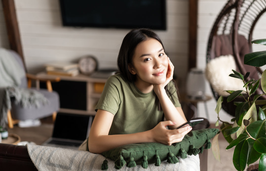 shutterstock_2103443501 A young woman in a green t-shirt sits indoors, leaning on a cushion, holding a smartphone, and looking at the camera. A laptop and home decor are visible in the background.