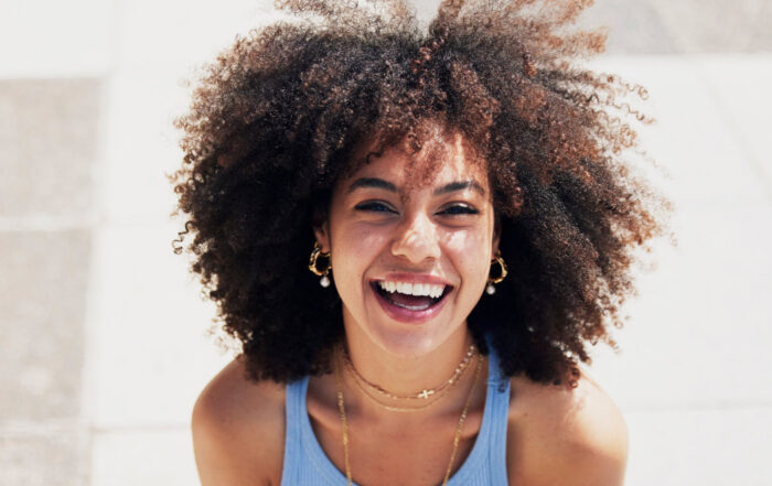 A person with curly hair, wearing a blue tank top and layered necklaces, smiles outdoors in bright sunlight.
