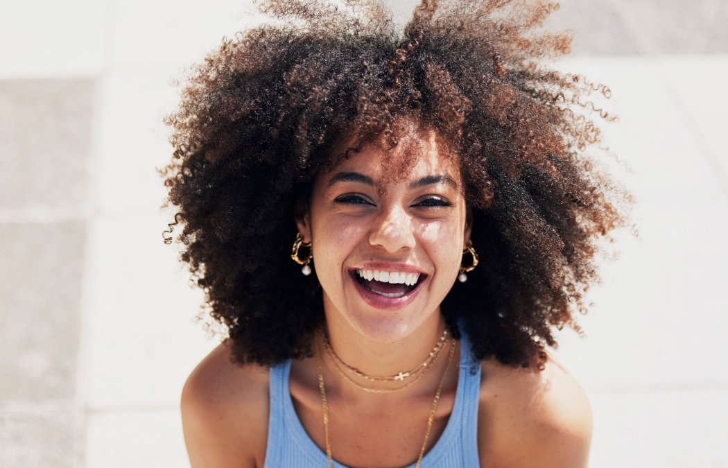 shutterstock_2260768875-1 A person with curly hair, wearing a blue tank top and layered necklaces, smiles outdoors in bright sunlight.